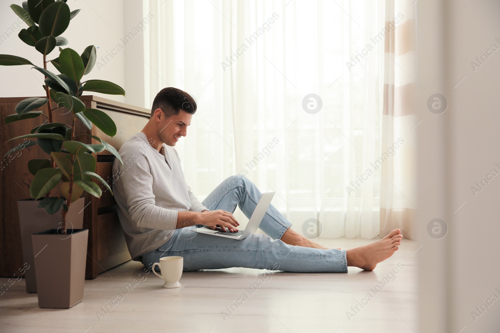 Photo of Man with cup of drink and laptop sitting on warm floor at home. Heating system