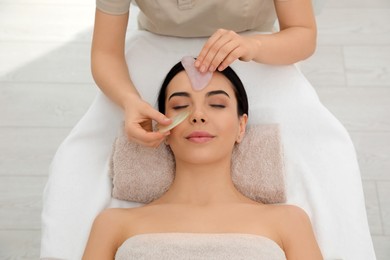 Photo of Young woman receiving facial massage with gua sha tool in beauty salon, top view