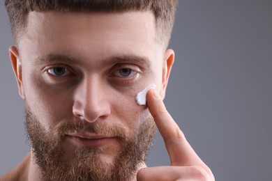 Handsome man applying moisturizing cream onto his face on grey background, space for text