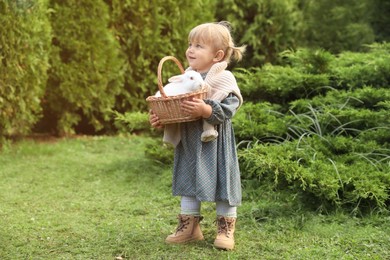 Happy little girl holding wicker basket with cute rabbit outdoors on sunny day. Space for text