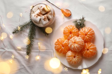 Photo of Peeled delicious ripe tangerines, festive lights and glass of drink with marshmallows on white bedsheet, flat lay