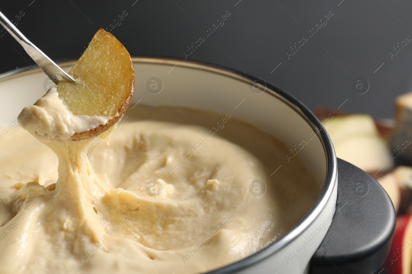 Photo of Dipping piece of potato into fondue pot with melted cheese on grey background, closeup