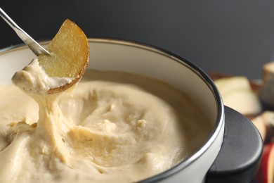 Photo of Dipping piece of potato into fondue pot with melted cheese on grey background, closeup