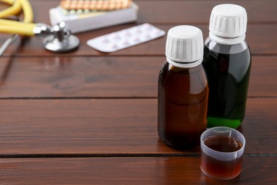 Photo of Bottles of syrup and measuring cup on wooden table, space for text. Cold medicine
