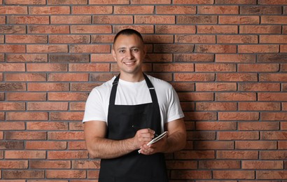 Portrait of happy young waiter with notebook near brick wall, space for text
