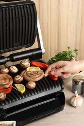 Photo of Woman cooking homemade sausages with mushrooms and bell pepper on electric grill at wooden table, closeup