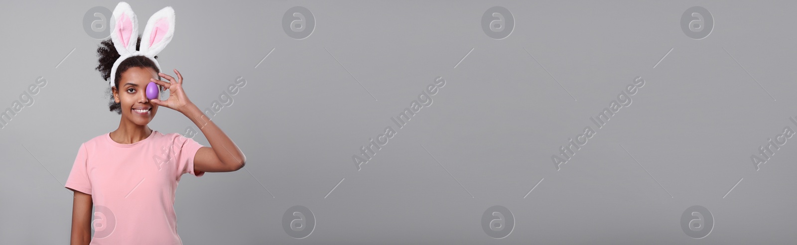 Image of Happy African American woman with bunny ears holding Easter egg near eye on light grey background, space for text. Banner design