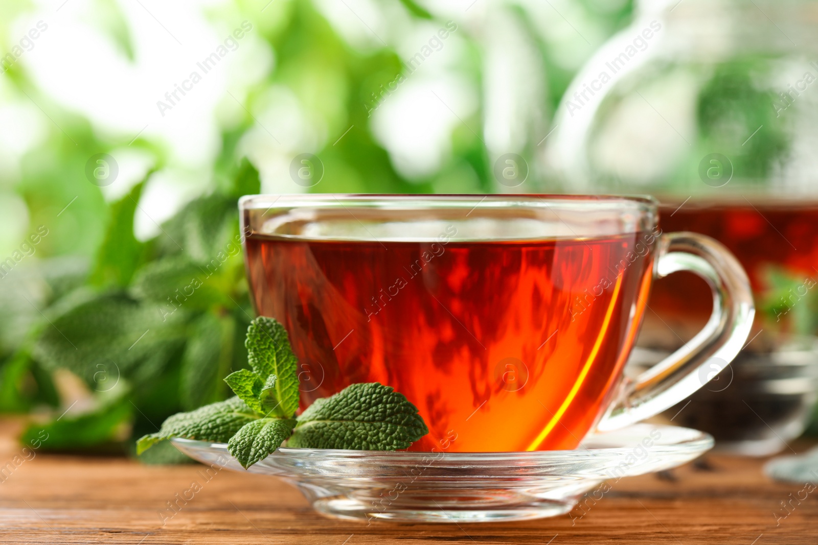 Photo of Glass cup of aromatic black tea with fresh mint on wooden table against blurred background, closeup