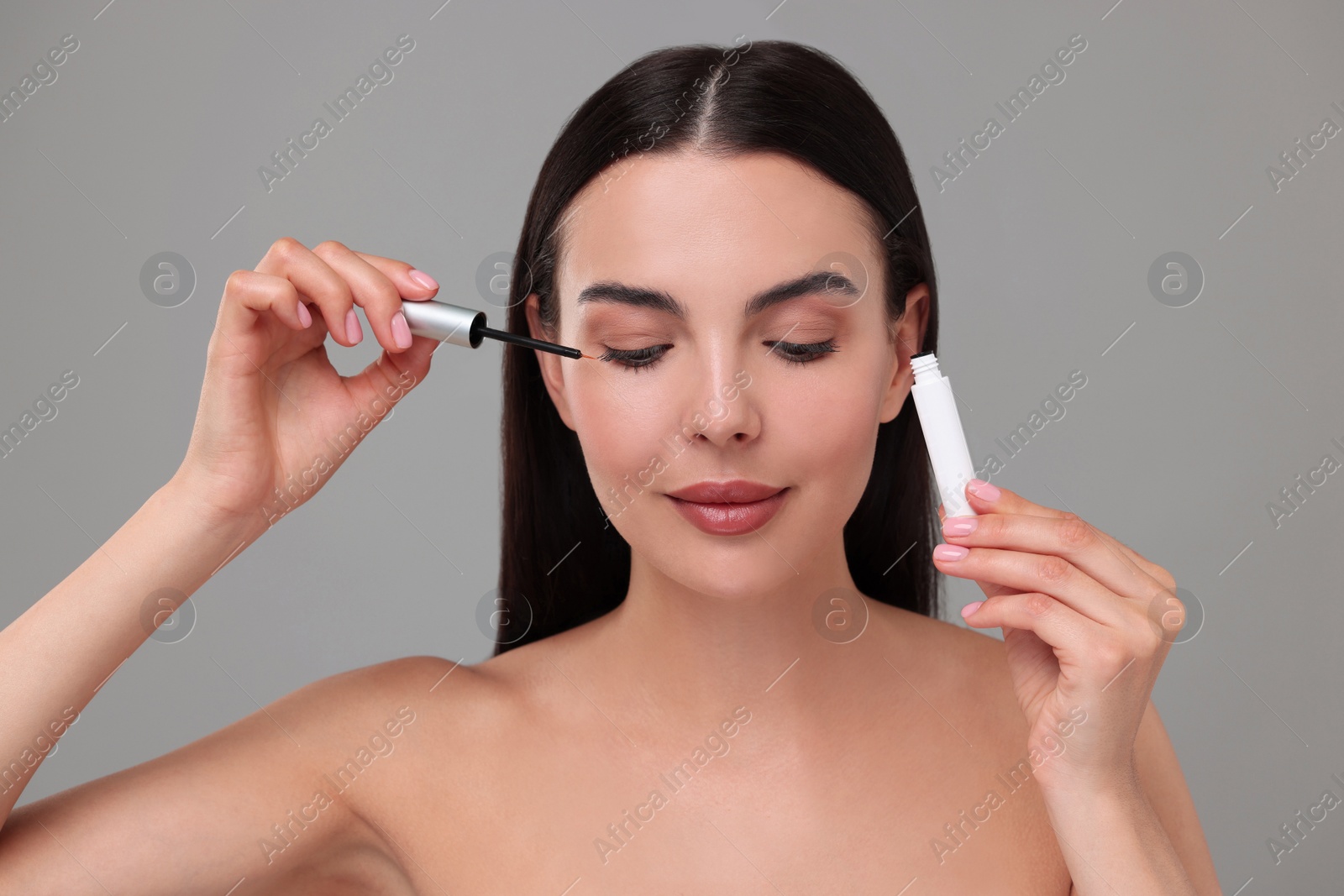 Photo of Beautiful woman applying serum onto her eyelashes on grey background. Cosmetic product