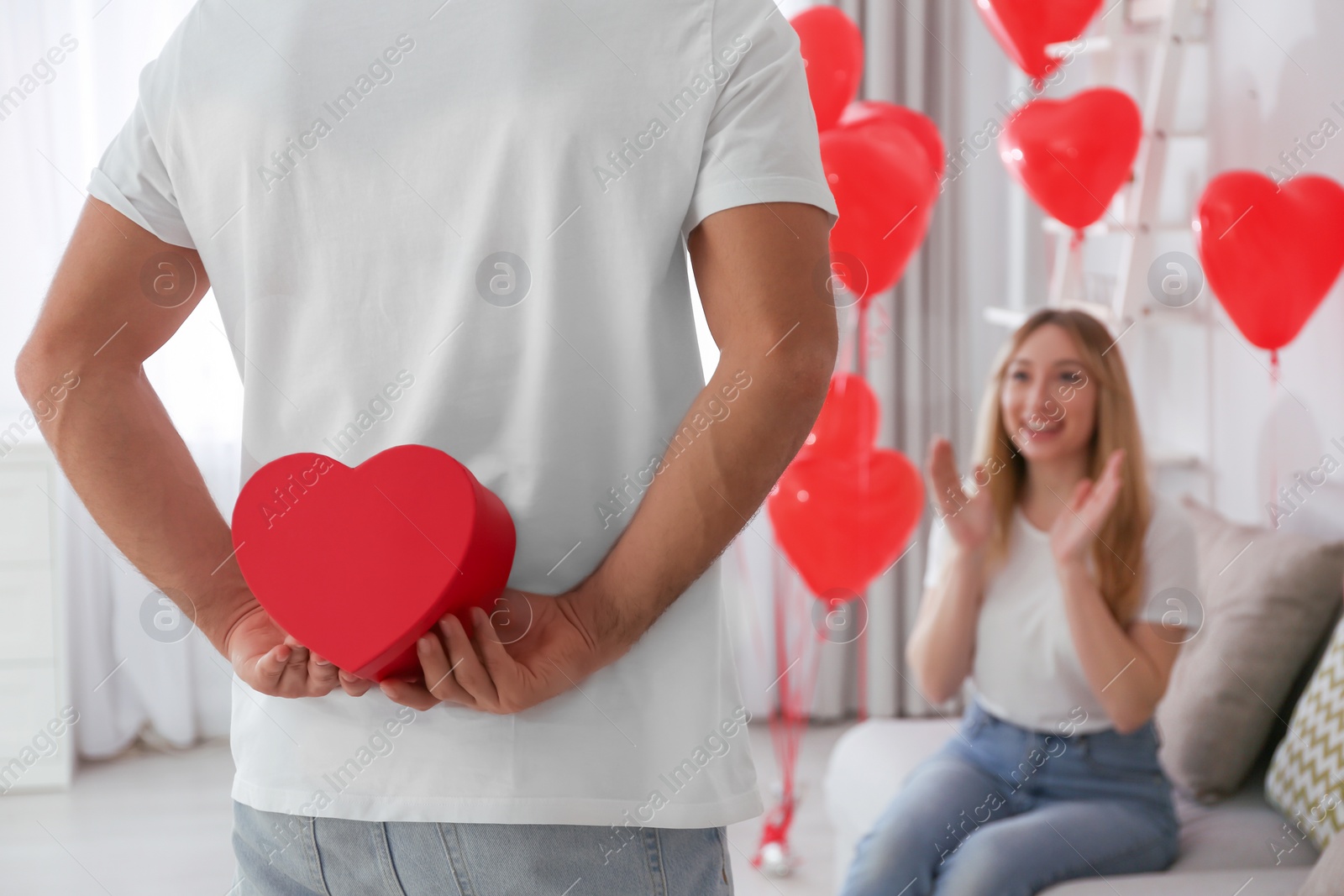 Photo of Man presenting gift to his girlfriend in room decorated with heart shaped balloons. Valentine's day celebration