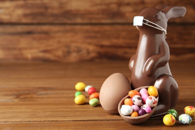 Photo of Chocolate bunny with protective mask, egg shaped candies and space for text on wooden table. Easter holiday during COVID-19 quarantine