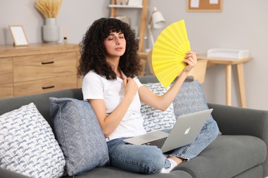 Photo of Young woman with laptop waving yellow hand fan to cool herself on sofa at home