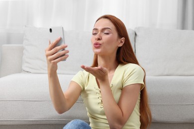 Photo of Happy young woman blowing kiss during video chat via smartphone at home. Long-distance relationship