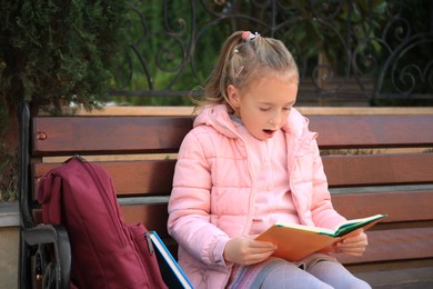 Cute little girl with backpack reading book on bench outdoors