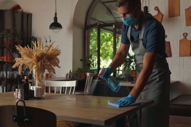 Photo of Waiter in mask and gloves disinfecting table at cafe
