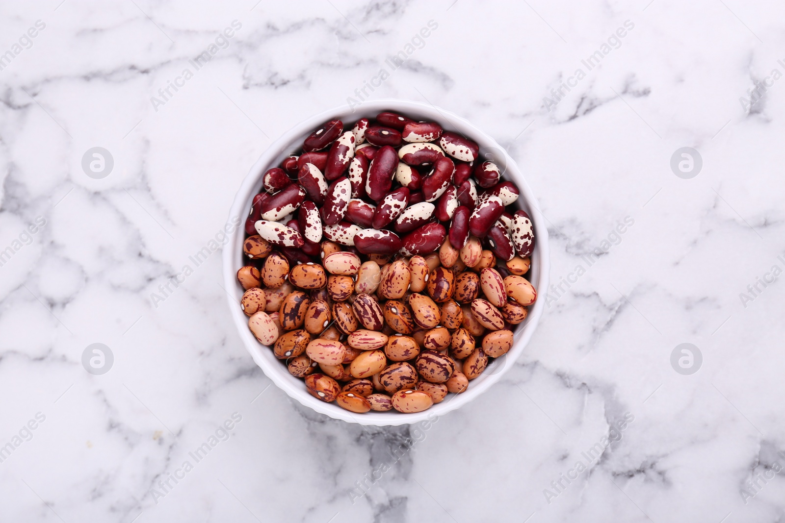 Photo of Different kinds of dry kidney beans in bowl on white marble table, top view