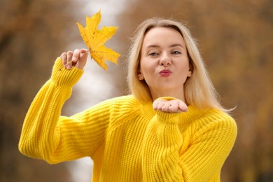 Beautiful woman with autumn leaf blowing kiss outdoors