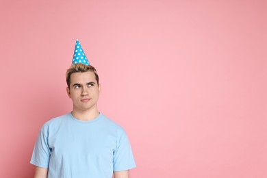 Sad young man with party hat on pink background, space for text