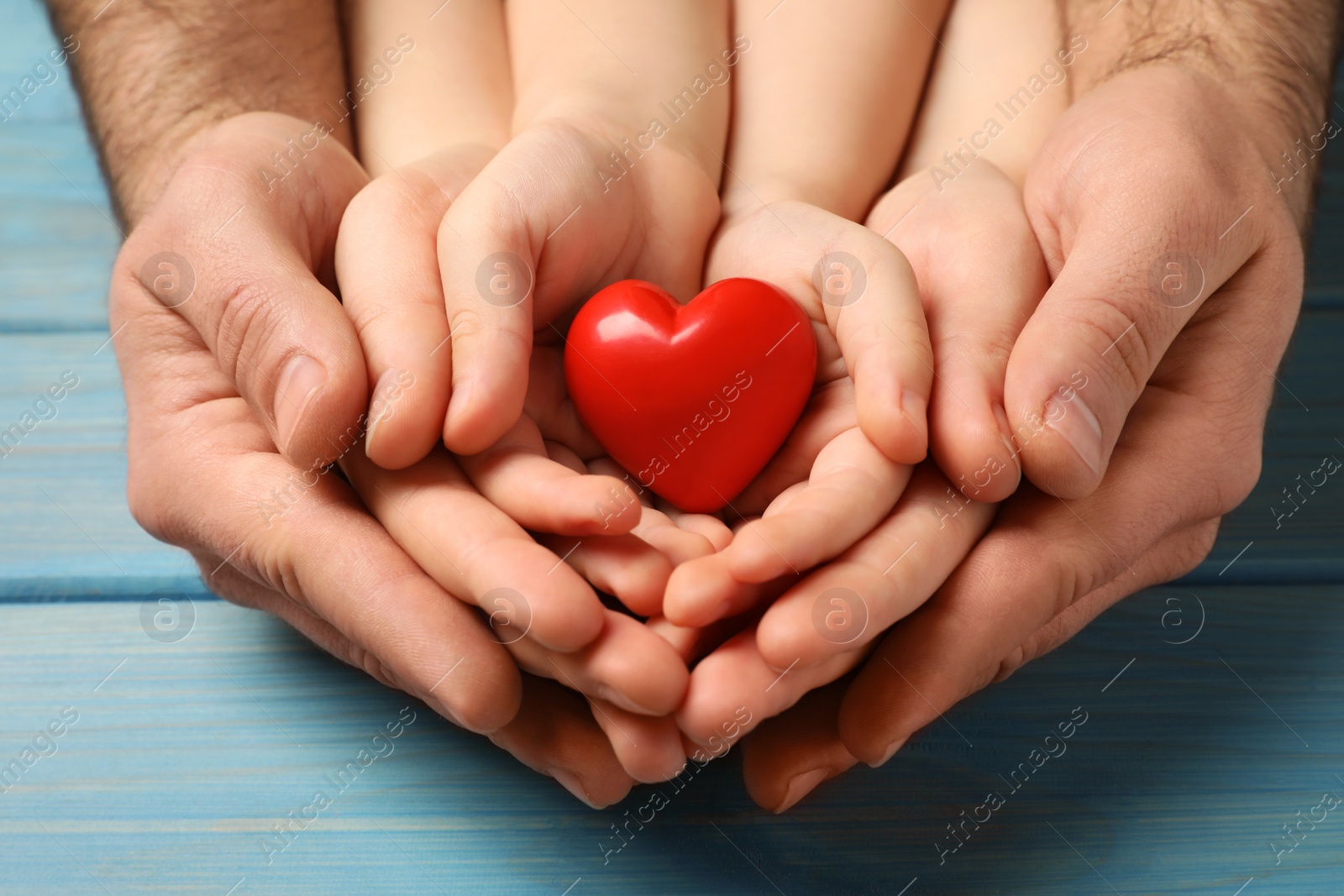 Photo of Parents and kid holding red heart in hands at light blue wooden table, closeup