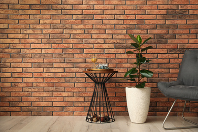 Stylish room interior with beautiful plant, chair and side table near brick wall. Space for text