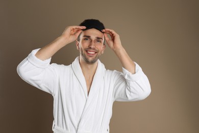 Photo of Happy young man in bathrobe and eye sleeping mask on brown background