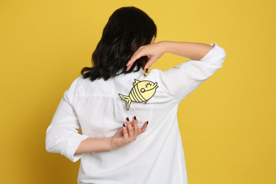Photo of Woman with paper fish on back against yellow background. April fool's day