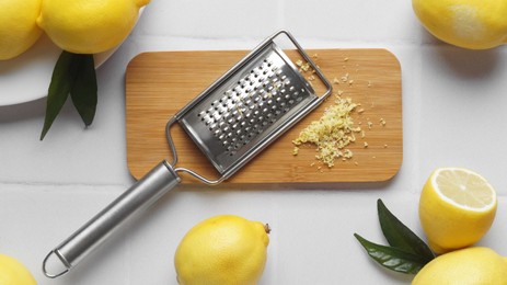 Photo of Wooden board with grater and fresh lemon zest on white tiled table, flat lay