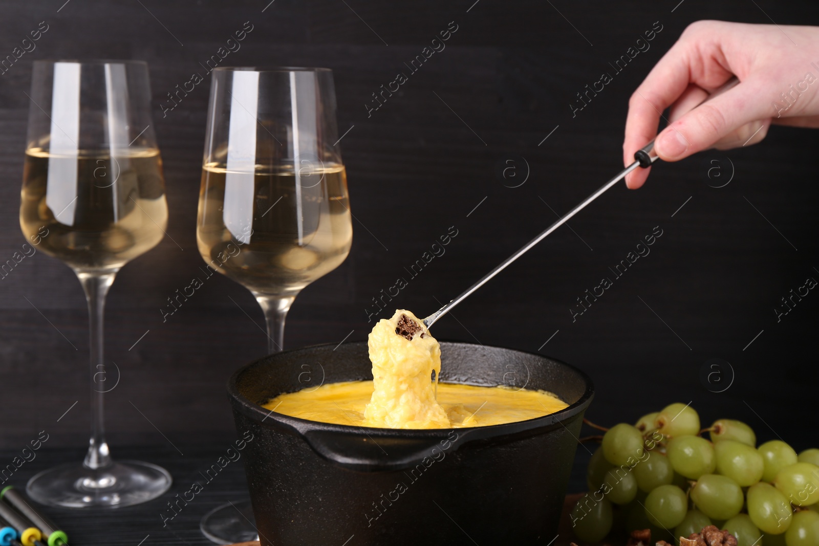 Photo of Woman dipping piece of bread into fondue pot with melted cheese at black wooden table, closeup