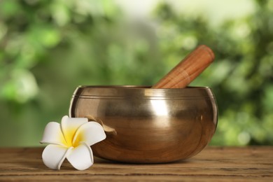 Photo of Golden singing bowl, mallet and flower on wooden table outdoors