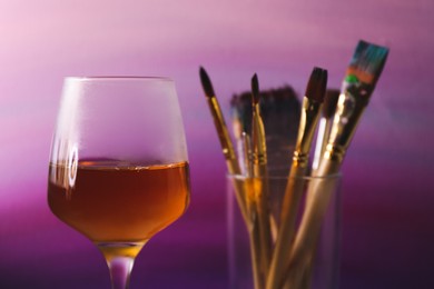 Photo of Glass of tasty wine, brushes with colorful paints near gradient canvas, closeup. Space for text