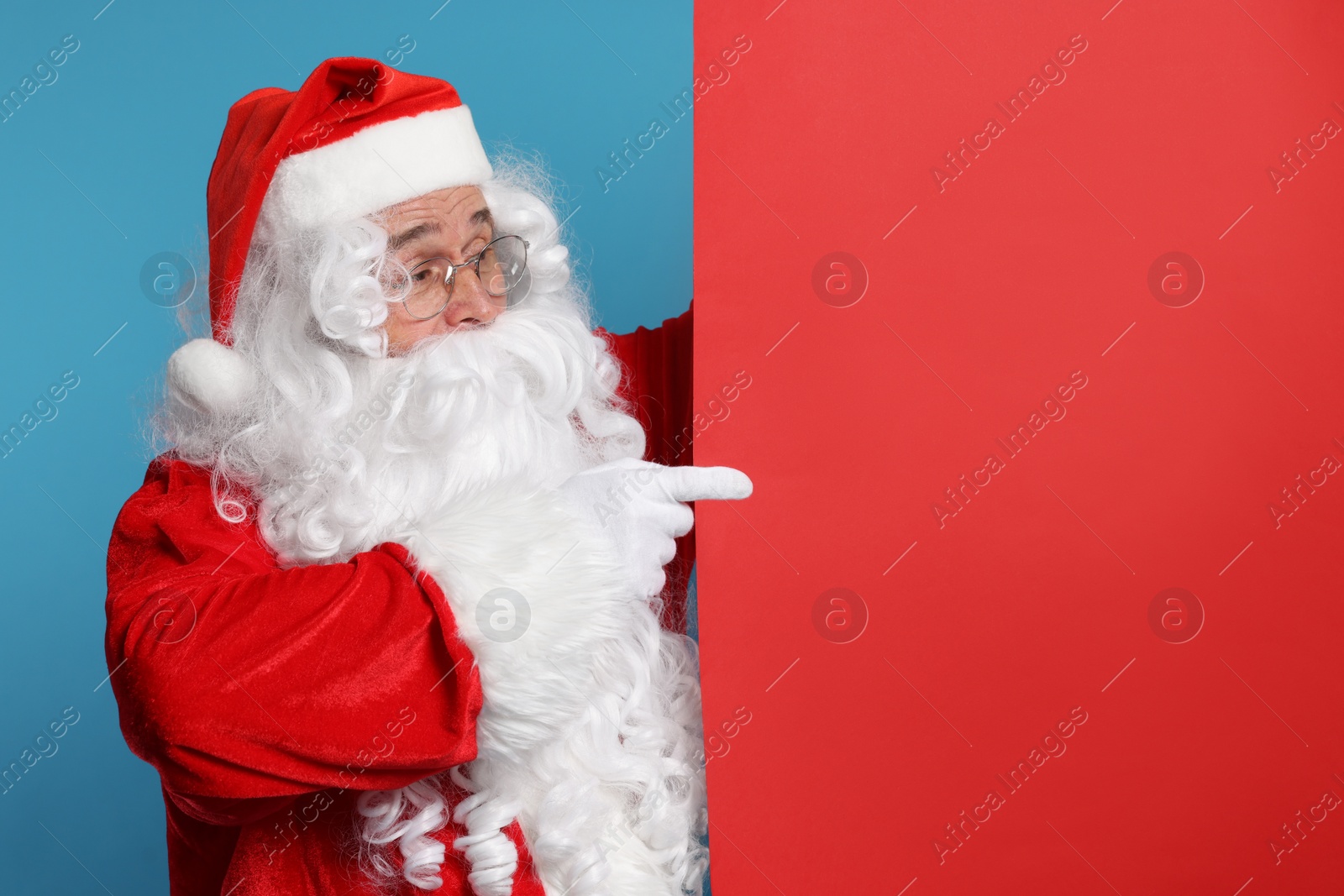 Photo of Santa Claus pointing at blank red poster on light blue background, space for text