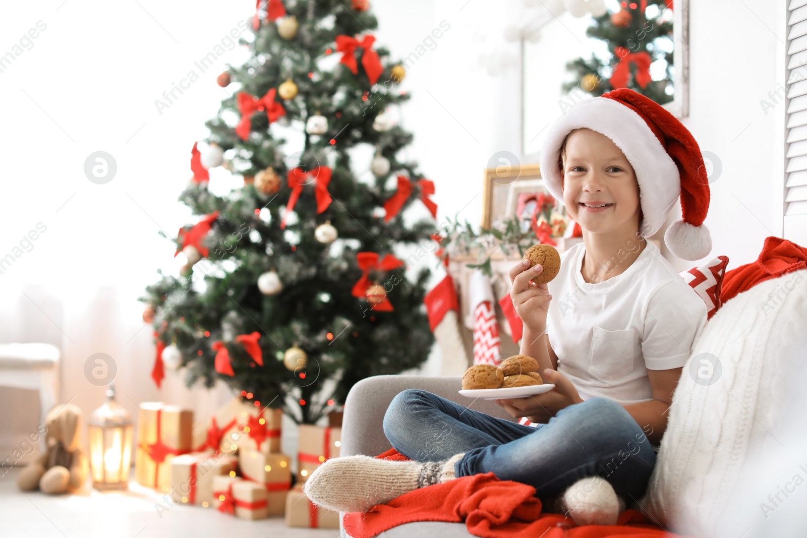 Photo of Cute little child in Santa hat eating Christmas cookies on sofa at home