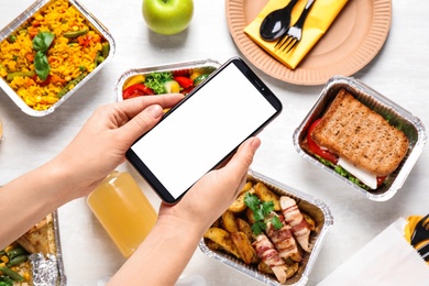 Photo of Top view of woman holding smartphone over white wooden table with lunchboxes,  mockup for design. Healthy food delivery