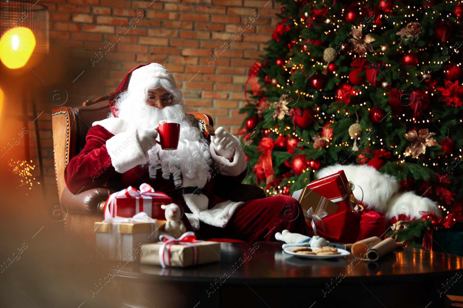 Photo of Santa Claus with hot drink and cookie near Christmas tree at home