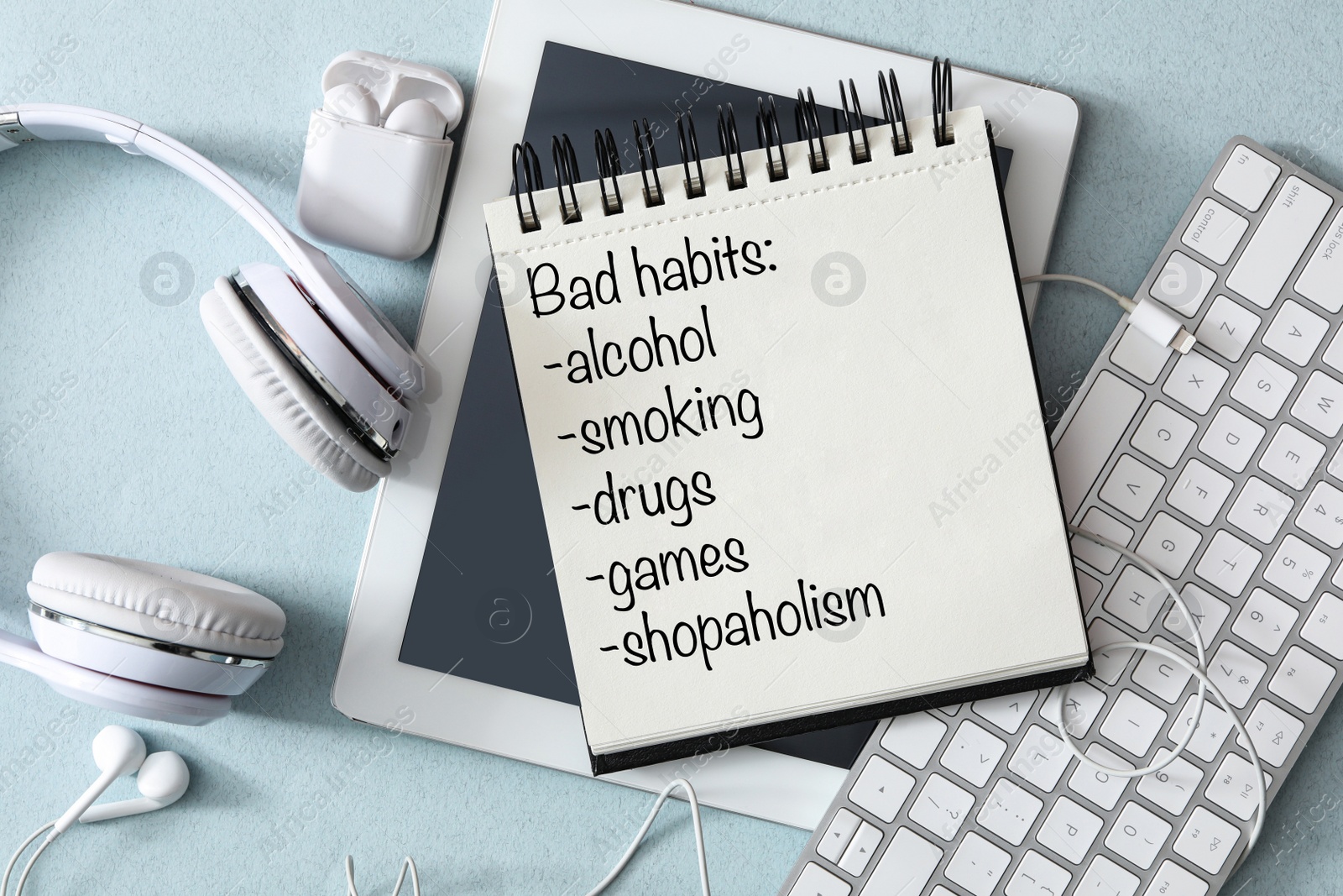 Image of Notebook with list of bad habits, different earphones, tablet and keyboard on light blue table, flat lay