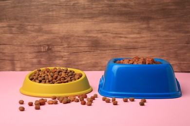 Photo of Dry and wet pet food in feeding bowls on pink table near wooden wall