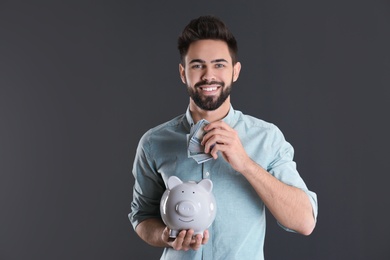 Happy young man putting money into piggy bank on grey background