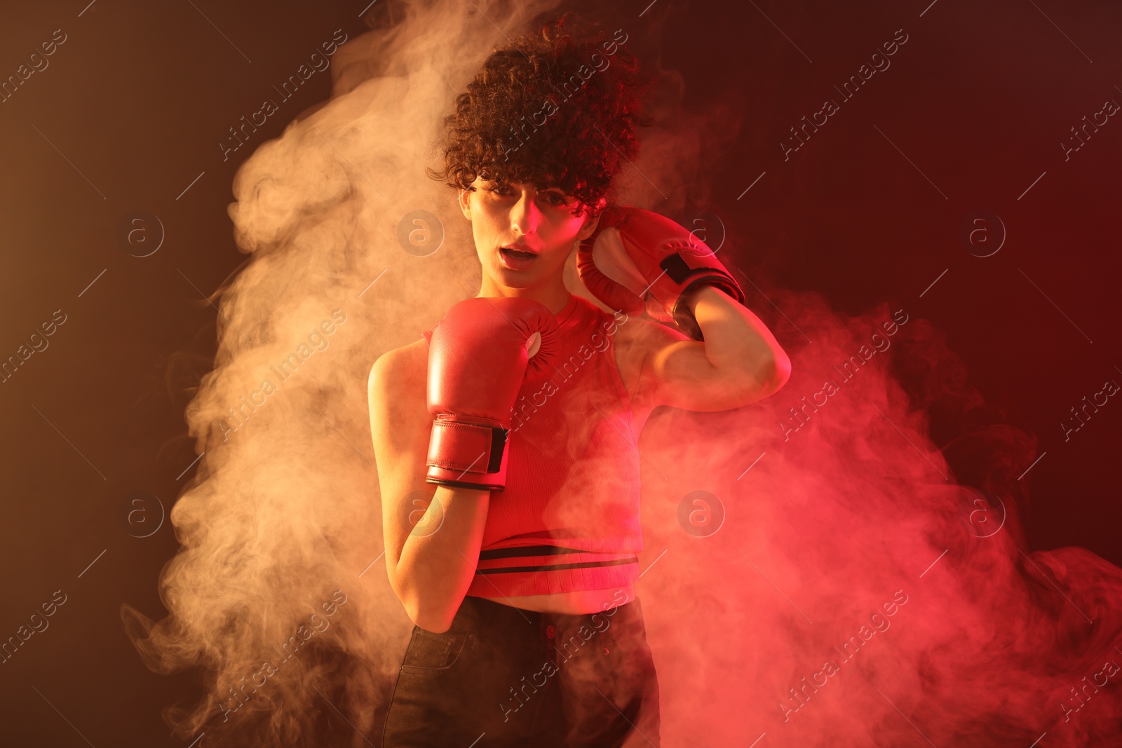 Photo of Beautiful young woman with boxing gloves on color background in neon lights and smoke