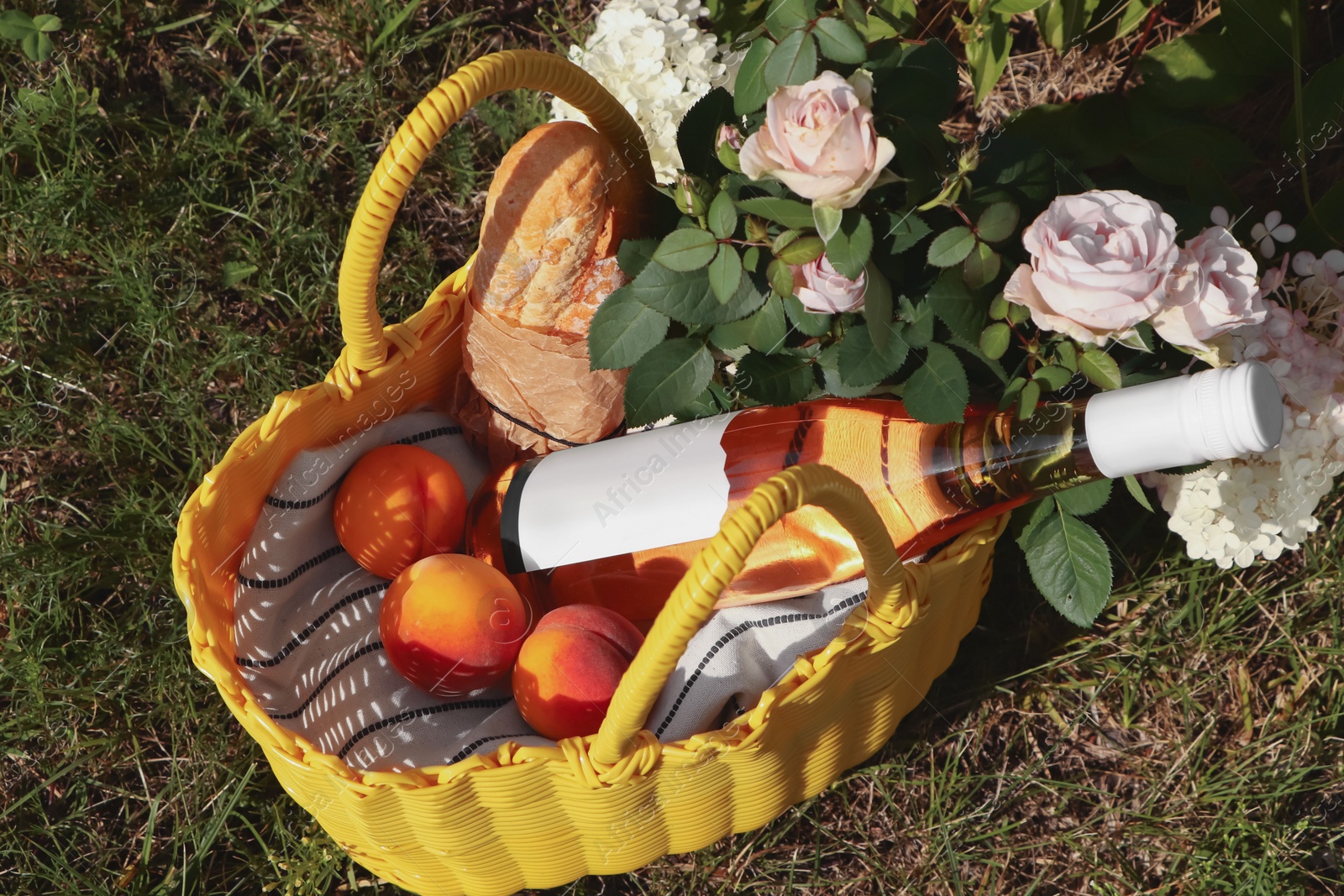 Photo of Yellow wicker bag with roses, peaches, baguette and wine outdoors, above view