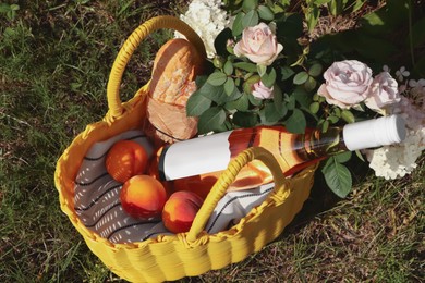 Photo of Yellow wicker bag with roses, peaches, baguette and wine outdoors, above view