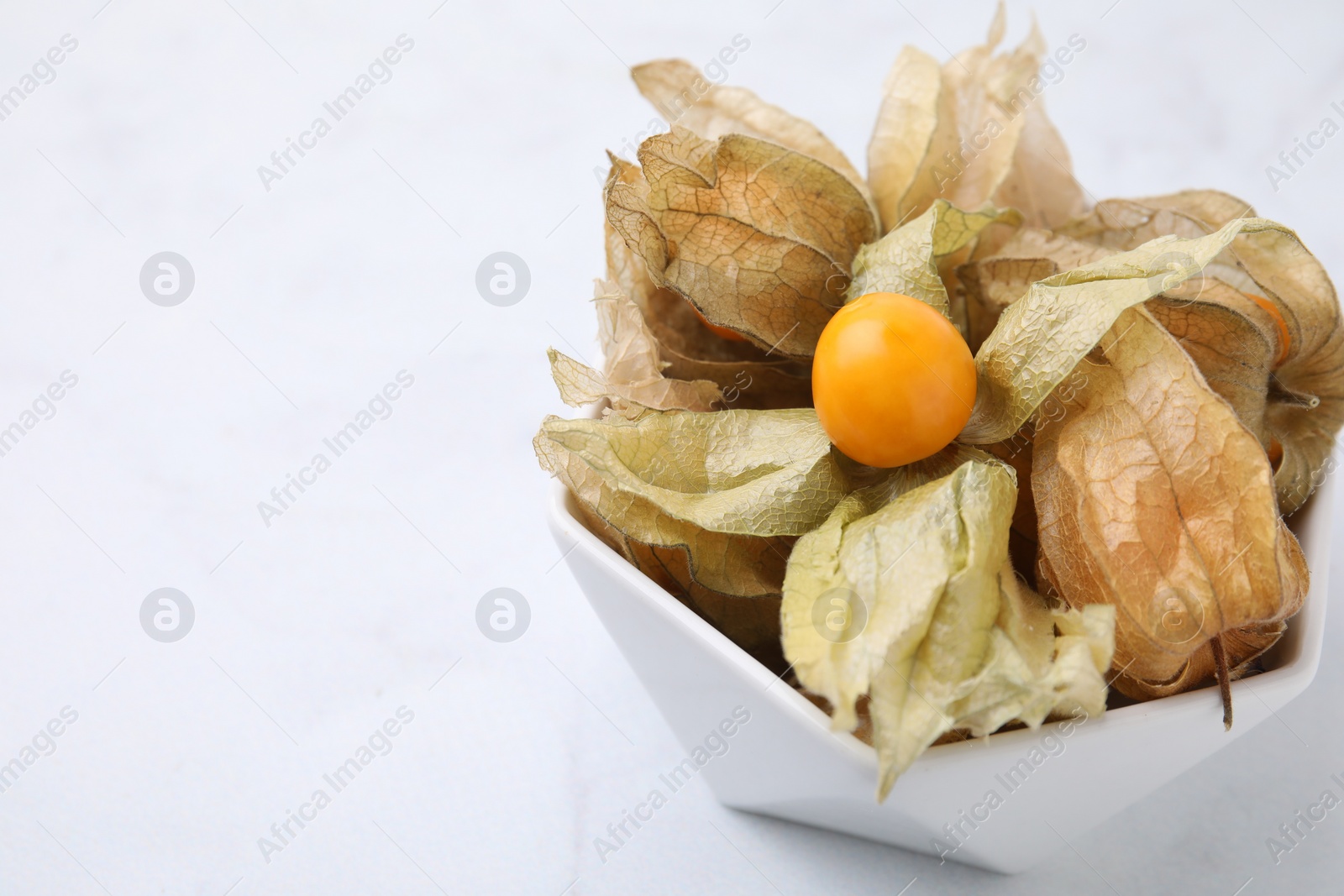 Photo of Ripe physalis fruits with calyxes in bowl on white marble table, closeup. Space for text