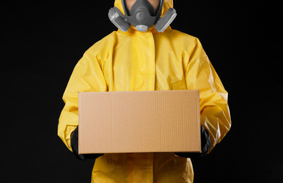 Photo of Man wearing chemical protective suit with cardboard box on black background, closeup. Prevention of virus spread