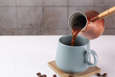 Photo of Pouring aromatic coffee from cezve into cup at white textured table, closeup. Space for text