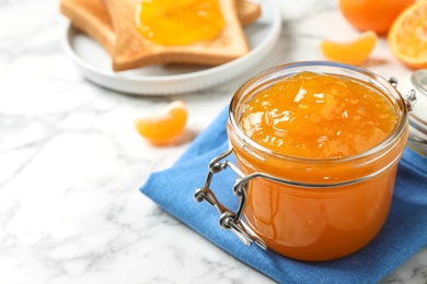 Tasty tangerine jam in glass jar on white marble table, space for text