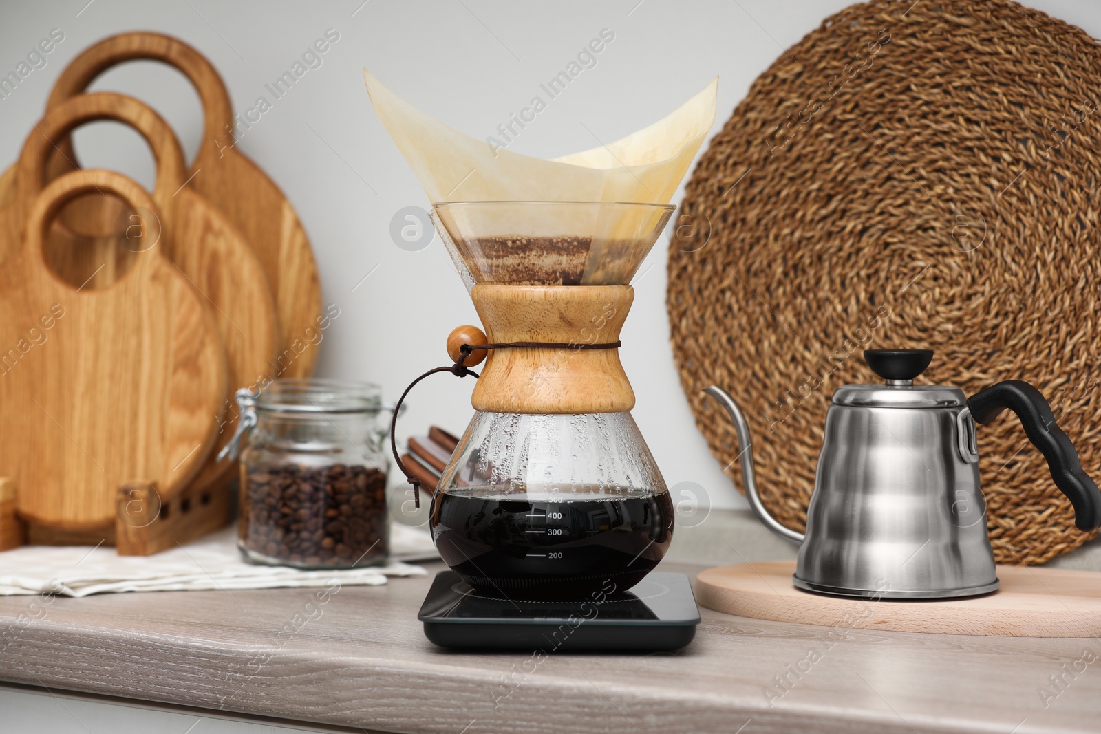 Photo of Making drip coffee. Glass chemex coffeemaker with paper filter, jar of beans and kettle on wooden countertop in kitchen