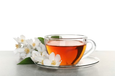 Glass cup of aromatic jasmine tea and fresh flowers on grey table against white background
