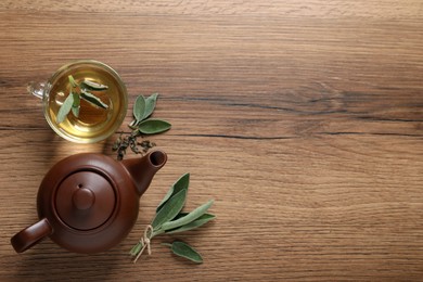 Photo of Cup of sage tea, green leaves and teapot on wooden table, flat lay. Space for text