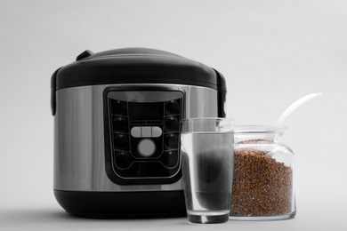 Photo of Modern electric multi cooker, buckwheat and glass of water on grey background