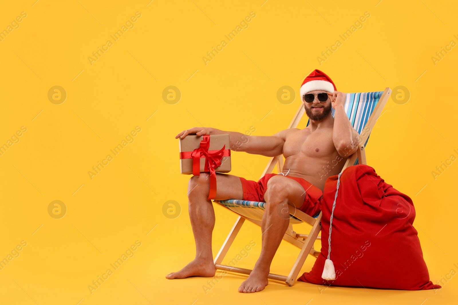 Photo of Muscular young man in Santa hat with deck chair, bag, sunglasses and Christmas gift box on orange background, space for text