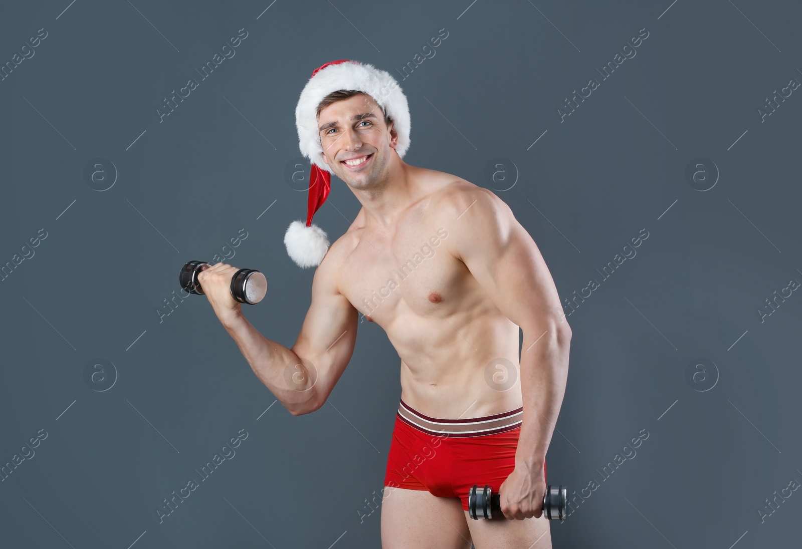 Photo of Young muscular man in Santa hat with dumbbells on gray background